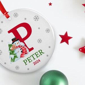 May include: White ceramic Christmas ornament with a red letter P, a snowman holding a "Merry Christmas" sign, and the name "Peter" with the year "2024" below.