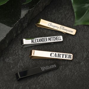 May include: Four personalized tie clips in different colors and finishes. The tie clips are engraved with the names "Gregory Thompson", "Alexander Mitchell", "Carter", and "Williams".