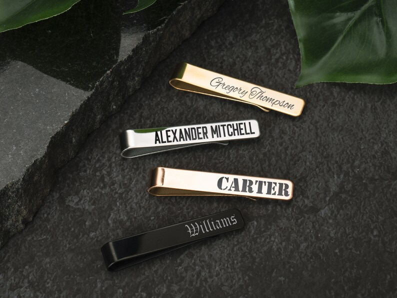 May include: Four personalized tie clips in different colors and finishes. The tie clips are engraved with the names "Gregory Thompson", "Alexander Mitchell", "Carter", and "Williams".