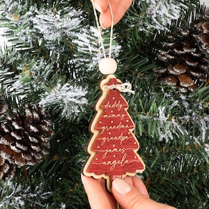 May include: A red wooden Christmas tree ornament featuring a layered design with white script text reading daddy, mommy, grandma, grandpa, james, and angela, accented by small stars and the year 2024 at the base, suspended by a string with a wooden bead.