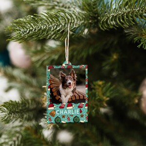 K&ouml;nnte beinhalten: Ein blaugr&uuml;ner Weihnachtsbaumschmuck mit einem Foto eines Hundes darin. Der Schmuck hat einen wei&szlig;en Rand und eine wei&szlig;e Schnur zum Aufh&auml;ngen. Der Schmuck ist mit einer rot-wei&szlig;en Weihnachtsm&uuml;tze, einer goldenen Glocke und einem roten Stern verziert. Der Name des Hundes, "CHARLIE", ist in Wei&szlig; auf den Schmuck gedruckt.