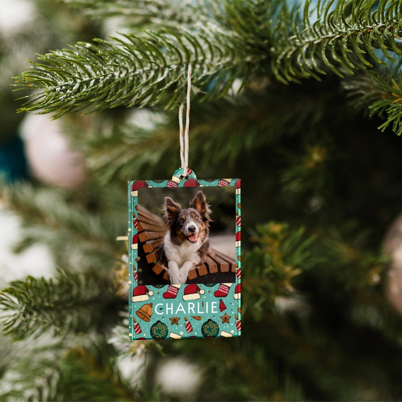 K&ouml;nnte beinhalten: Ein blaugr&uuml;ner Weihnachtsbaumschmuck mit einem Foto eines Hundes darin. Der Schmuck hat einen wei&szlig;en Rand und eine wei&szlig;e Schnur zum Aufh&auml;ngen. Der Schmuck ist mit einer rot-wei&szlig;en Weihnachtsm&uuml;tze, einer goldenen Glocke und einem roten Stern verziert. Der Name des Hundes, "CHARLIE", ist in Wei&szlig; auf den Schmuck gedruckt.