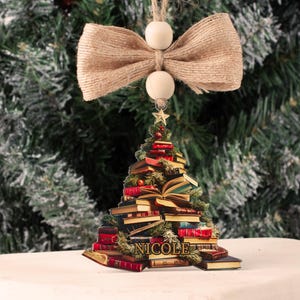 May include: A wooden Christmas tree ornament shaped like a stack of books with a star on top. The ornament is decorated with a burlap bow and the name "NICOLE" is written on the base.