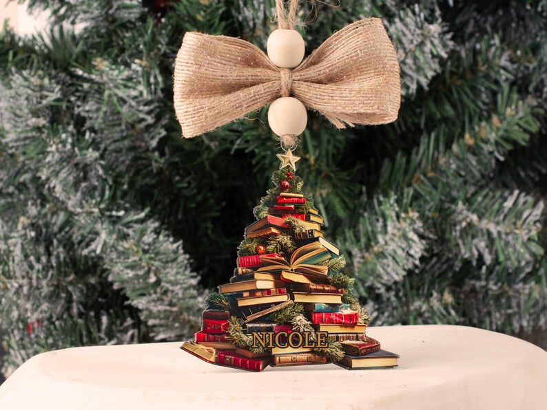 May include: A wooden Christmas tree ornament shaped like a stack of books with a star on top. The ornament is decorated with a burlap bow and the name "NICOLE" is written on the base.