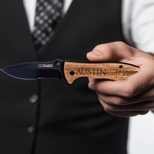 May include: A black pocket knife with a wooden handle. The handle is engraved with the name "Austin" and the text "Groomsman 21.01.2023".