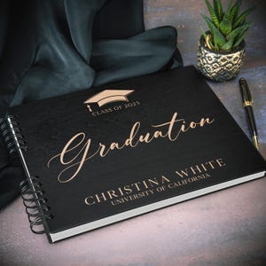 May include: Black graduation guest book with a gold inscription that reads "Class of 2025 Graduation Christina White University of California".