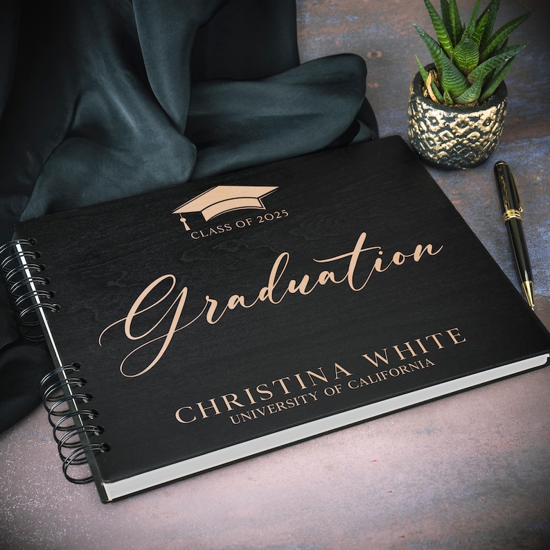 May include: Black graduation guest book with a gold inscription that reads "Class of 2025 Graduation Christina White University of California".
