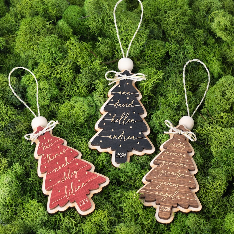 May include: Three Christmas tree-shaped wooden ornaments in red, black, and brown. Each ornament features white script names, small star accents, and the year 2024 engraved at the base. They hang from white strings attached to wooden beads.