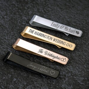 May include: Four personalized tie clips in different metal finishes: silver, gold, rose gold, and black. The tie clips are engraved with names and symbols like hearts, infinity signs, and wedding rings.