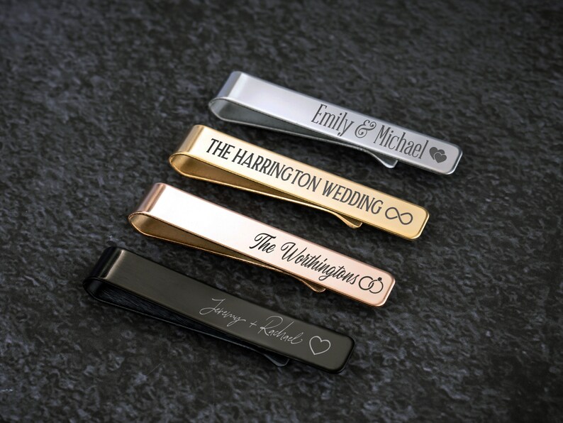 May include: Four personalized tie clips in different metal finishes: silver, gold, rose gold, and black. The tie clips are engraved with names and symbols like hearts, infinity signs, and wedding rings.