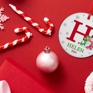 May include: White round Christmas ornament with a red letter H, the name HELEN and the year 2024. The ornament features a snowman with a green hat and scarf, holding a gift.