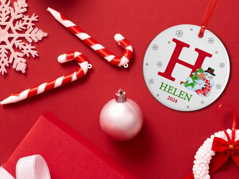 May include: White round Christmas ornament with a red letter H, the name HELEN and the year 2024. The ornament features a snowman with a green hat and scarf, holding a gift.