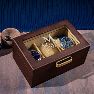 May include: A dark brown wooden watch box with a glass top and a gold handle. The box has a beige interior with slots for watches and a personalized inscription that reads "M Moss".