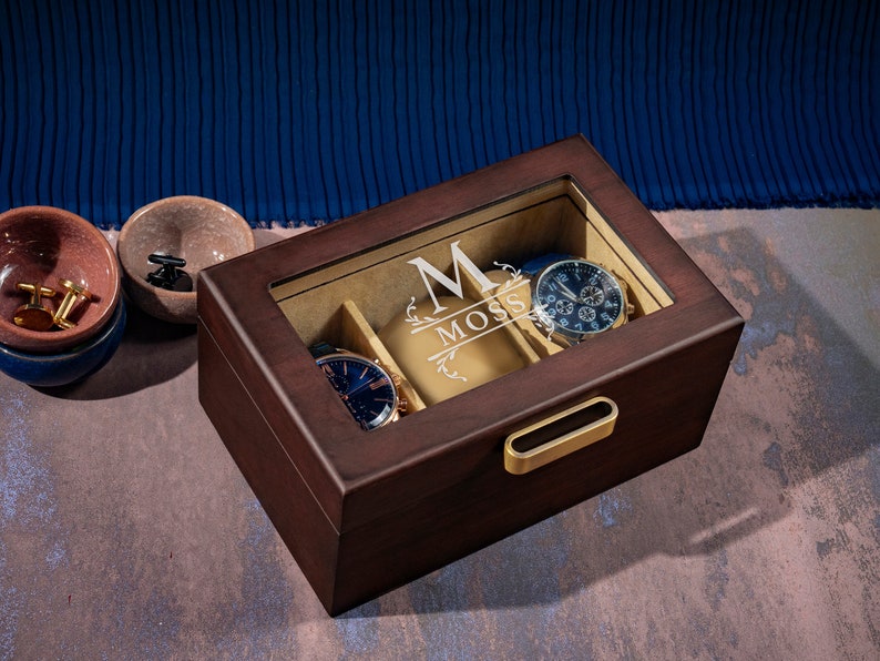 May include: A dark brown wooden watch box with a glass top and a gold handle. The box has a beige interior with slots for watches and a personalized inscription that reads "M Moss".