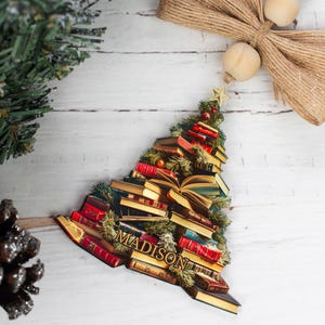 May include: A Christmas tree ornament shaped like a stack of books with the name "MADISON" written on the front. The ornament is decorated with red and gold accents and has a green base.