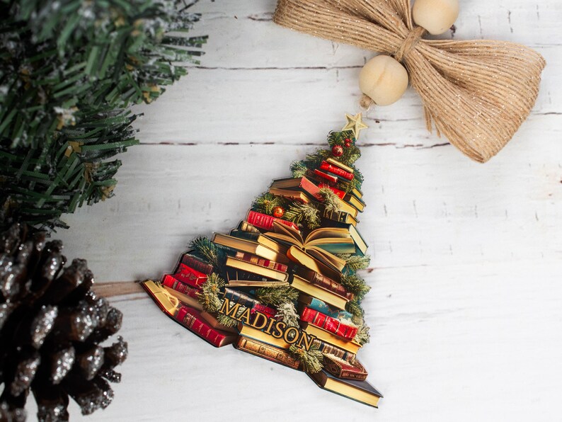 May include: A Christmas tree ornament shaped like a stack of books with the name "MADISON" written on the front. The ornament is decorated with red and gold accents and has a green base.