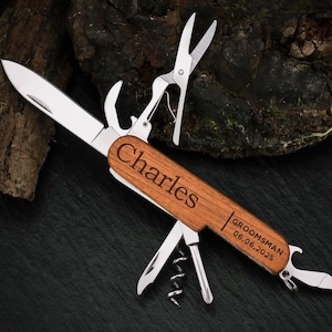 May include: A multi-tool pocket knife with a wooden handle engraved with the name "Charles" and the text "Groomsman 06.06.2025". The knife has a variety of tools, including a blade, a bottle opener, a corkscrew, and a pair of scissors.