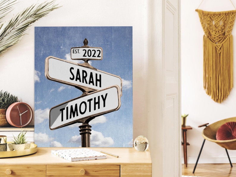Op de afbeelding: Een gepersonaliseerd straatnaambord met de namen "Sarah" en "Timothy" en de tekst "Est. 2022". Het bord is tegen een blauwe lucht met witte wolken.