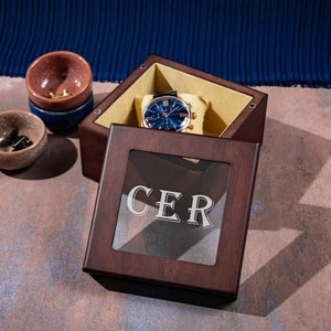 May include: A dark brown wooden box with a glass top. The box has a gold watch inside and the word "CER" engraved on the glass.