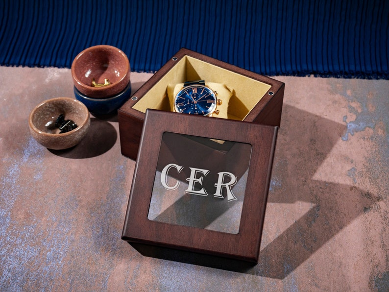 May include: A dark brown wooden box with a glass top. The box has a gold watch inside and the word "CER" engraved on the glass.
