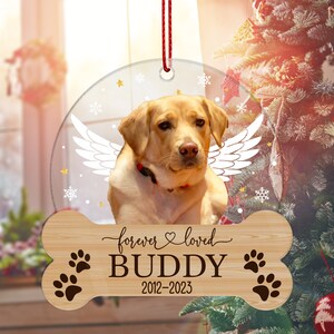 May include: A circular clear ornament featuring a yellow Labrador retriever with white angel wings, suspended by a red string. Below, a bone-shaped wooden plaque displays the text forever loved, the name BUDDY, and the years 2012-2023.