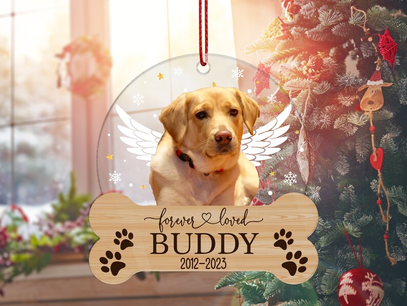 May include: A circular clear ornament featuring a yellow Labrador retriever with white angel wings, suspended by a red string. Below, a bone-shaped wooden plaque displays the text forever loved, the name BUDDY, and the years 2012-2023.