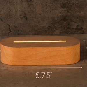 May include: A rectangular wooden LED light base, 5.75 inches long and 1.38 inches high, with a glowing horizontal light strip. The light is made of light-colored wood and has rounded edges. A white power cord extends from the base.