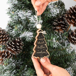 May include: A black wooden Christmas tree ornament with layered edges, featuring the names ana, david, hellen, and andrea in white script, small white stars, and the year 2024 at the base, suspended by a white string with a wooden bead.