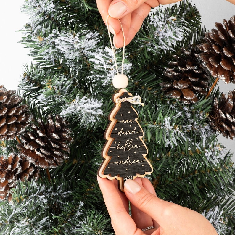 May include: A black wooden Christmas tree ornament with layered edges, featuring the names ana, david, hellen, and andrea in white script, small white stars, and the year 2024 at the base, suspended by a white string with a wooden bead.