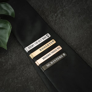 May include: Four tie clips with engraved names and titles. The tie clips are silver, gold, rose gold, and black.