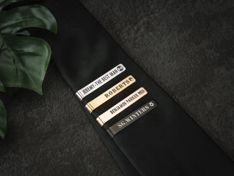 May include: Four tie clips with engraved names and titles. The tie clips are silver, gold, rose gold, and black.