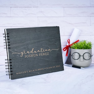 May include: A dark gray wooden scrapbook with a spiral binding. The cover is engraved with the words "graduation" and "Joshua Perez" in a rose gold font. The text "Midtown High School - Class of 2023" is also engraved on the cover.