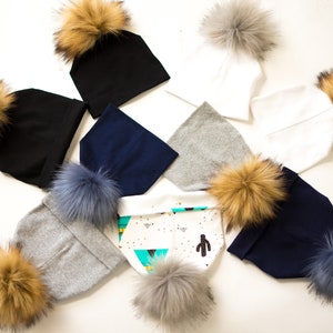 May include: Assortment of knit beanies with faux fur pom poms in various colors, including black, white, gray, and blue. The beanies are arranged on a white surface.