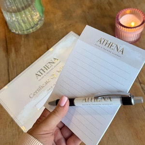 Könnte beinhalten: Ein Notizblock, ein Stift und ein Zertifikat der Athena Salon & Training Academy werden auf einer Holzoberfläche präsentiert. Der Stift und der Notizblock tragen das Athena-Logo. Eine brennende Kerze und eine Vase mit Grünzeug befinden sich im Hintergrund.