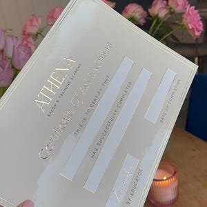May include: A certificate of achievement from Athena Salon & Training Academy. The certificate is a light cream color with gold lettering and a gold border. The text reads "Certificate of Achievement" and "This is to certify that:" with spaces for completion details.