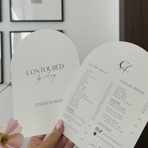 Może przedstawiać: W ręku trzymane są dwa białe, łukowate menu zabiegów. Lewe menu zawiera napis "Contoured by Amy" i "menu zabiegów". Prawe menu zawiera listę usług usuwania owłosienia laserem i drenażu limfatycznego.