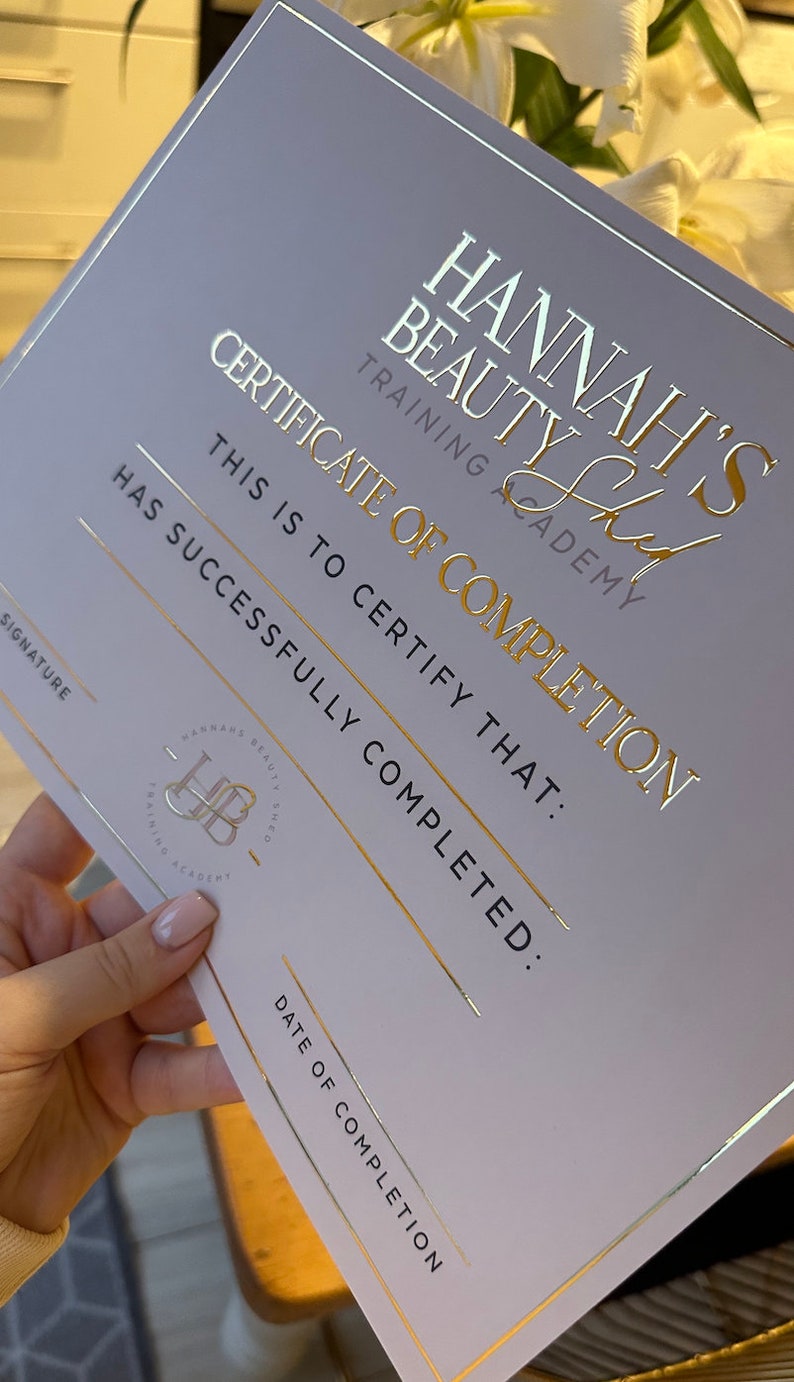 May include: A certificate from Hannah's Beauty Training Academy. The certificate is light gray with gold foil lettering and accents. The text reads "Certificate of Completion" and includes the academy's logo.