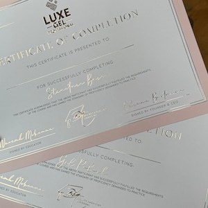 May include: Two pink and white certificates with gold lettering. The top certificate reads "LUXE -GEL- PROFESSIONAL CERTIFICATE OF COMPLETION" and is presented to "Stenituen Bes". The bottom certificate reads "CERTIFICATE OF COMPLETION" and is presented to "Gol P.lul". Both certificates are signed by "Oksana Britmane, SIGNED BY FOUNDER & CEO".