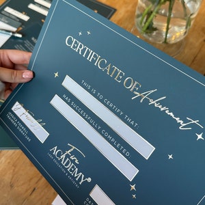 May include: A Certificate of Achievement from Fire Academy, in a dark teal color with gold lettering. The certificate includes spaces for the recipient's name, and the date of course completion. The certificate is held in front of a wooden table.