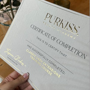 May include: A certificate of completion from Purkiss Beauty Academy for a Classic Lash Extensions Training Course. The certificate has gold lettering and a gold border. The certificate is held by a person.