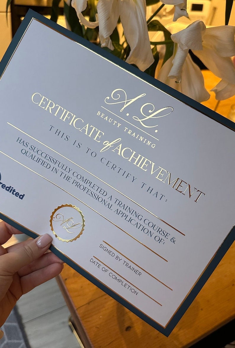 May include: A certificate of achievement from A.L. Beauty Training. The certificate has gold foil lettering and a gold seal. The text reads "Certificate of Achievement" and certifies completion of a training course. The certificate has a dark green border.