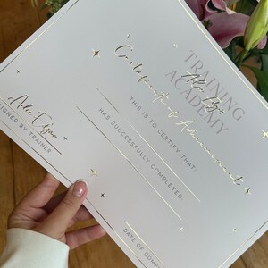 May include: A white certificate with gold foil lettering and accents. The certificate reads "Training Academy Certificate of Achievement" and includes lines for completion details. The certificate is held near pink lilies.