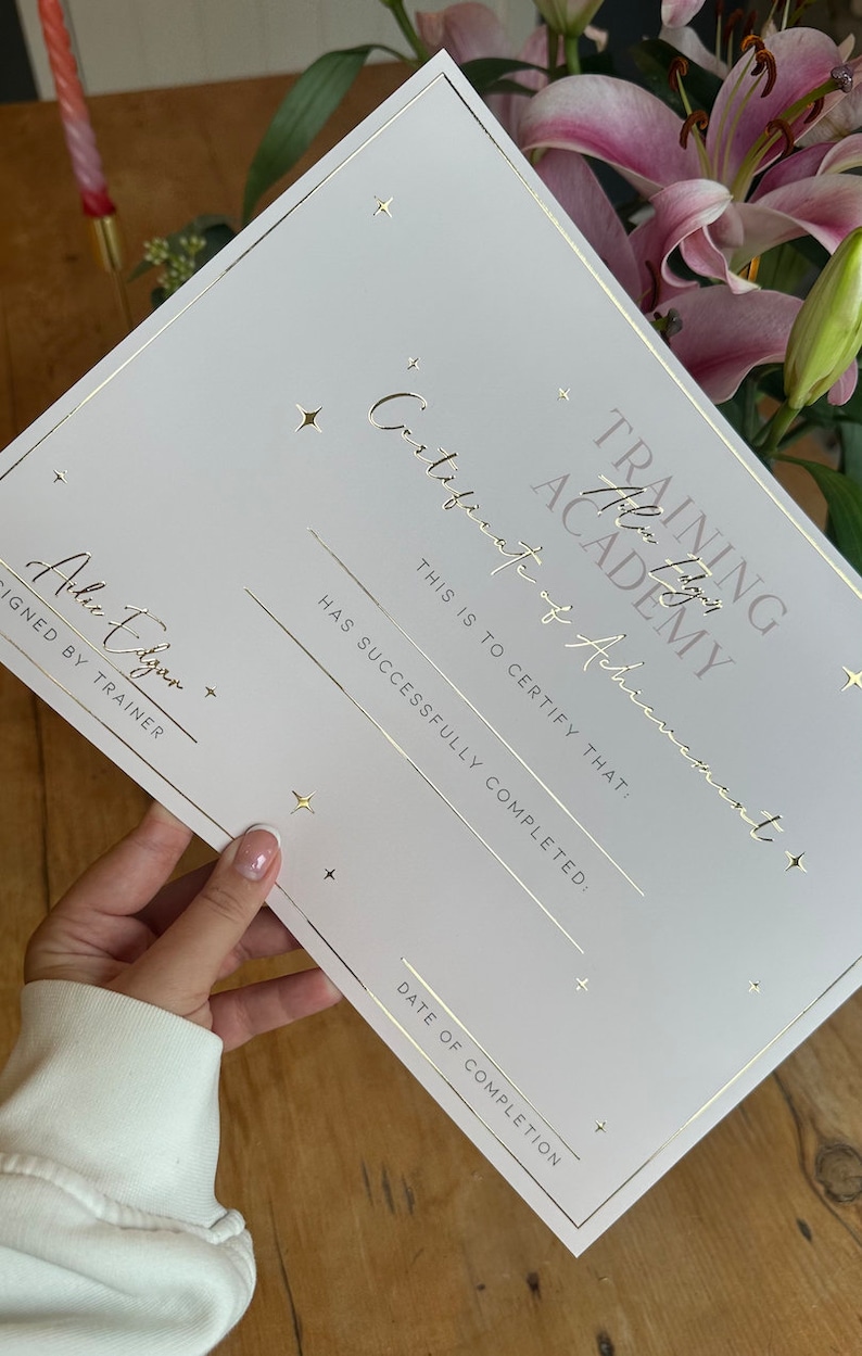 May include: A white certificate with gold foil lettering and accents. The certificate reads "Training Academy Certificate of Achievement" and includes lines for completion details. The certificate is held near pink lilies.