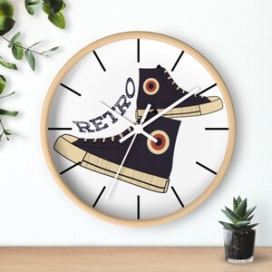 May include: A round wall clock with a light wood frame. The clock face features a graphic of two black high-top sneakers with the word "RETRO" in blue text. The sneakers have tan soles and red and tan circular accents.