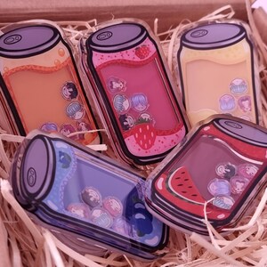 May include: Five colorful acrylic charms shaped like soda cans, each filled with small, decorative elements. The cans feature various fruit designs, including strawberry and watermelon, and are set in a wooden box with straw.