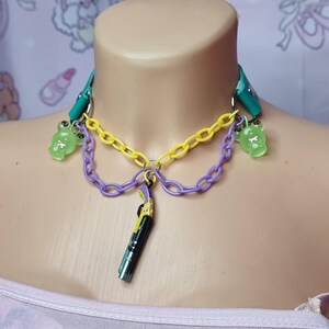 May include: A multi-layered necklace with a green, yellow, and purple chain. The necklace has a small black and yellow charm hanging from it. The necklace is decorated with green gummy bear charms.