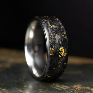 May include: A black carbon fiber ring with gold flecks. The ring is polished and has a smooth finish.