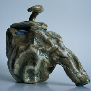 Puede incluir: Una escultura de cerámica con una forma orgánica texturizada. La escultura está esmaltada en tonos de marrón, blanco y azul. Tiene un pico y una tapa con un asa curva única.