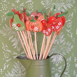 Könnte beinhalten: In einer grünen Metallkanne befinden sich Holzdekorationen von Früchten und Gemüse an Stäben, darunter Äpfel, Kürbisse und Birnen. Die Ausschnitte sind in Orange, Rot und Grün bemalt und mit weißen Blumenmustern verziert.