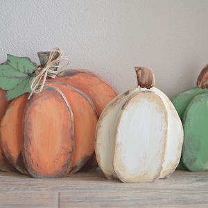 Könnte beinhalten: Drei dekorative Holzkürbisse in Orange, Weiß und Grün. Der große orangefarbene Kürbis hat ein grünes Blatt und eine Schleife aus Jutegarn am Stiel. Jeder Kürbis ist bemalt und weist an den Rändern dunkle Akzente auf, was für Tiefe sorgt.